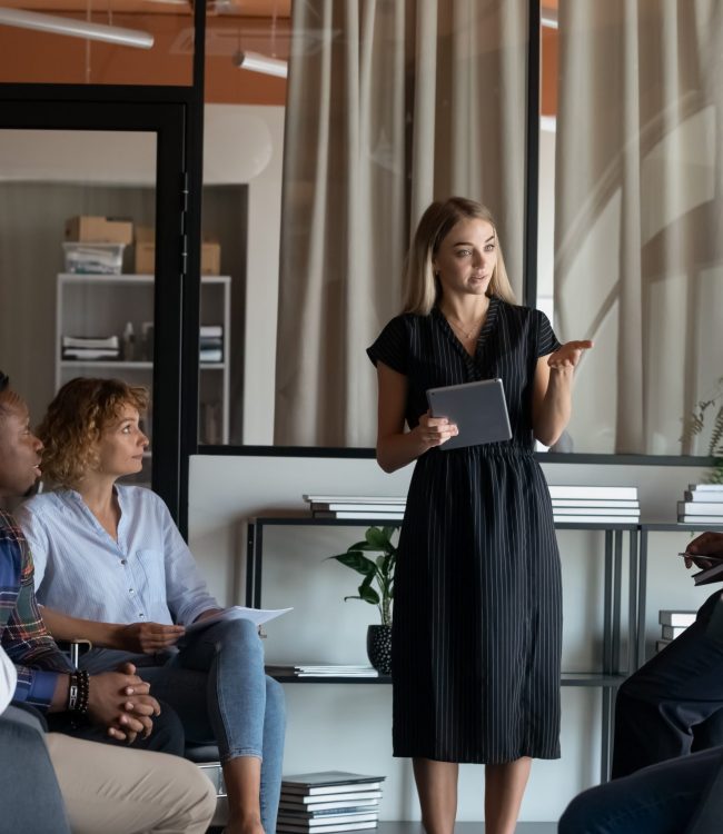 Confident businesswoman mentor holding tablet leading corporate meeting briefing with diverse employees in modern office boardroom, colleagues coworkers listening to executive, sitting in circle
